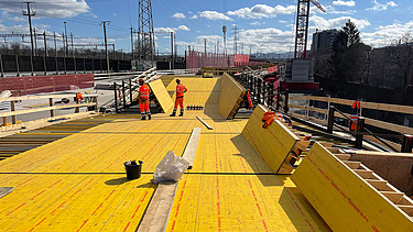  Ein Kran der STRABAG hebt nachts ein langes Brückenteil über Bahngleise. Die Baustelle ist hell erleuchtet.
