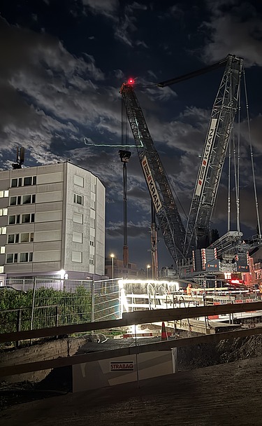 Nachtaufnahme einer STRABAG-Baustelle. Ein grosser Kran steht beleuchtet neben einem Wohnhaus unter dem klaren Nachthimmel.