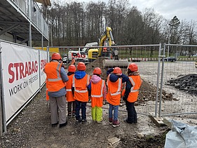 Eine Gruppe von Schulkindern in orangefarbenen Schutzwesten und Helmen steht vor einem Zaun und beobachtet einen Bagger bei Bauarbeiten auf einer Baustelle in Kloten.