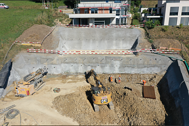 Ein Bagger befindet sich in einer grossen Baugrube. Mehrere Erdschichten sind deutlich zu erkennen, die Baustelle ist mit einem Absperrband gesichert. Im Hintergrund stehen moderne Wohnhäuser.