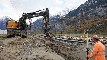 Ein Bagger hebt auf einer Baustelle Erdmaterial aus, während ein Arbeiter in orangefarbener Schutzkleidung daneben steht und den Vorgang beobachtet. Im Hintergrund liegen Gleise sowie die typischen Absperrelemente, dahinter eine steile Berglandschaft mit Wald und Fels.