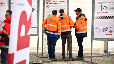Mehrere Personen in orangefarbener Schutzkleidung stehen vor Informationstafeln einer Baustelle. Im Vordergrund ist unscharf eine STRABAG-Fahne zu sehen.