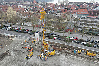 Auf einer Baustelle steht ein grosses gelbes Bohrgerät, das tiefe Löcher für Grossbohrpfähle in den Boden bohrt. Mehrere Arbeiter und Baumaschinen sind im Einsatz.