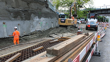 Baustelle an einer Strasse mit Nagelwand, mehreren Baumaschinen und einem Arbeiter in oranger Kleidung an einem bewölkten Tag.