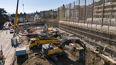 Rüschlikon Bahnhof Süd – STRABAG Tiefbau und Spezialtiefbau Drohnenaufnahme der Baugrube von vorne, zu sehen sind mehrere Bagger, einen Kran im Hintergrund sowie die Bahnlinie die direkt neben der Grube langläuft