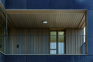 Ein Balkon mit einer Holzdecke und Wänden aus Holz. Die Fassade besteht aus senkrechten Holzlatten.