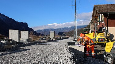 Mehrere Arbeiter in orangefarbener Schutzkleidung stehen auf einer frisch geschotterten Strasse. Eine gelbe Baumaschine steht direkt daneben. Rechts sind Wohnhäuser zu sehen.