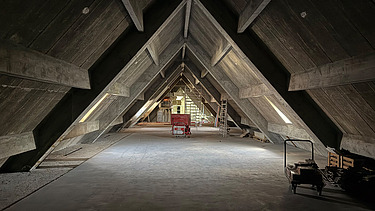 Historischer Dachstuhl mit markanter, dreieckiger Holzkonstruktion in A-Form. In der Mitte des Raumes steht ein roter Baustellenwagen, rechts eine Leiter und Baumaterial. Durch seitliche Fensteröffnungen fällt etwas Tageslicht ins Innere. Der Raum befindet sich noch im Umbau.