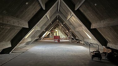 Historischer Dachstuhl mit markanter, dreieckiger Holzkonstruktion in A-Form. In der Mitte des Raumes steht ein roter Baustellenwagen, rechts eine Leiter und Baumaterial. Durch seitliche Fensteröffnungen fällt etwas Tageslicht ins Innere. Der Raum befindet sich noch im Umbau.