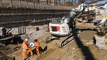 Drohnenaufnahme der Baugrube von vorne, zu sehen sind mehrere Bagger, im Vordergrund Bauarbeiter bei Vermessungen die zusammen in einem Kreis stehen