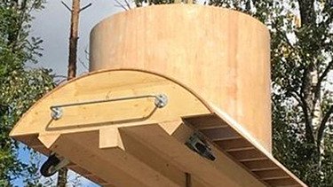 Ein Kran der STRABAG hebt an einem sonnigen Tag ein grosses Brückenelement. Im Hintergrund stehen mehrere Personen auf der Baustelle, neben einem Wohngebäude.