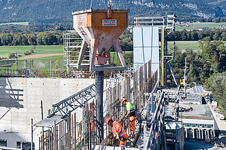 Bauarbeiter giessen Beton in eine hohe Schalung. Ein Betontrichter hängt am Kran, im Hintergrund ist die Landschaft mit Feldern und Bergen zu sehen.