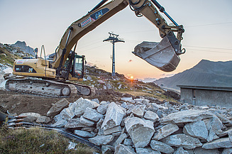 Ein Bagger von STRABAG bewegt grosse Steine auf einer Baustelle im Gebirge bei Sonnenaufgang.