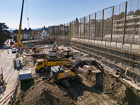 Drohnenaufnahme der Baugrube von vorne, zu sehen sind mehrere Bagger, einen Kran im Hintergrund sowie die Bahnlinie die direkt neben der Grube langläuft