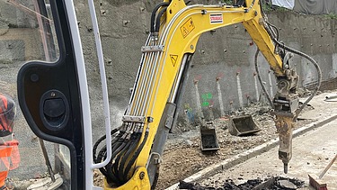 Eine kompakte Baumaschine spitzt Asphalt in einem Graben auf; daneben sind Baustellenmaterialien und Absperrungen sichtbar