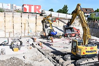 Mehrere Baufahrzeuge in einer Baugrube bei der Arbeit. Im Hintergrund ein weisser STRABAG Container mit der Aufschrift STR in grossen roten Buchstaben.