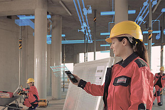Eine Frau mit Schutzhelm und Tablet steht auf einer Baustelle und überprüft digitale Baupläne. Im Hintergrund arbeiten weitere Personen.