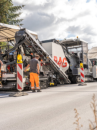STRABAG - Mitarbeiter und Maschinen bei Strassenbauarbeiten; eine Asphaltkolonne trägt frischen Asphalt auf eine Strasse auf.