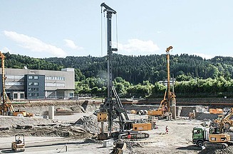 Auf einer Baustelle im Spezialtiefbau arbeiten mehrere Baumaschinen, darunter ein grosses Bohrgerät und Bagger. Im Hintergrund sind ein Industriegebäude und bewaldete Hügel zu sehen.