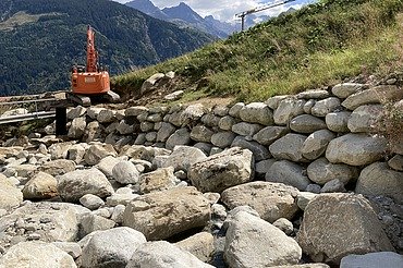 Schutz vor Naturgefahren - STRABAG Ingenieurbau STRABAG-Bagger baut Steinschutzwand in den Bergen am Aclettbach bei Disentis. Im Hintergrund ist eine Berglandschaft zu sehen.