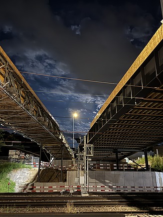 Die Aufnahme zeigt zwei grosse, eingerüstete Brückenteile, die sich links und rechts über den Gleisen erheben. In der Mitte führt der Blick in die beleuchtete Baustelle, darüber ein dramatischer Nachthimmel mit Wolken.