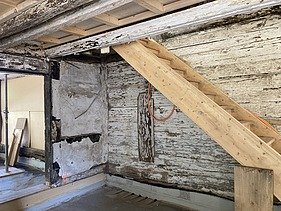 Ein Treppe aus hellem neuen Holz im Adlisberghof mit alten, abblätternden Wänden und Holzbalken.