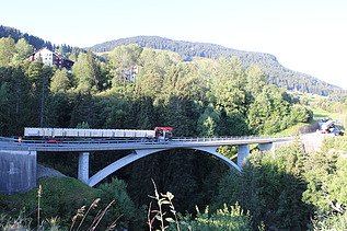 Ein langer Lastwagen fährt über eine hohe Brücke, die durch ein dicht bewaldetes Tal führt. Im Hintergrund sieht man steile, grüne Hänge und blauen Himmel.