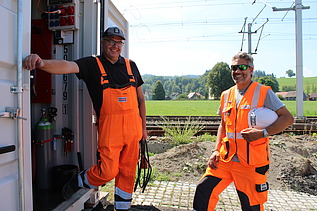 Zwei Männer in orangefarbener Bauausrüstung stehen lachend vor einem geöffneten Container auf der Baustelle.