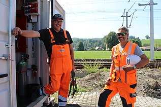 Zwei Männer in Bauausrüstung, lächelnd vor dem Container