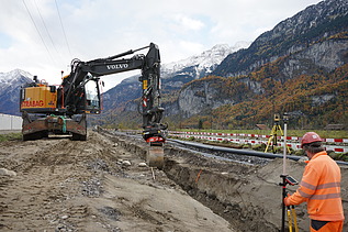 Ein Bagger hebt auf einer Baustelle Erdmaterial aus, während ein Arbeiter in orangefarbener Schutzkleidung daneben steht und den Vorgang beobachtet. Im Hintergrund liegen Gleise sowie die typischen Absperrelemente, dahinter eine steile Berglandschaft mit Wald und Fels.