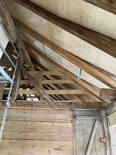 Blick in einen Dachstuhl mit Holzbalken und Verkleidung, links ist ein Gerüst zu sehen. Eine Kombination aus altem vorhandenem Holz mit neuem Holzelementen. 