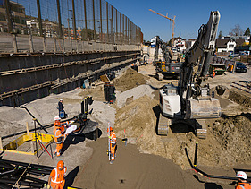 Drohnenaufnahme der Baugrube von vorne, zu sehen sind mehrere Bagger, im Vordergrund Bauarbeiter bei Vermessungen