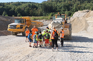 STRABAG Mitarbeiter mit 13 Kindern und zwei Erwachsenen in Kiesgruppe mit Pneulader und Dumper