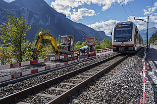 Ein Zug fährt auf einem Gleis durch eine Baustelle, rechts und links stehen Baumaschinen und Absperrungen. Im Hintergrund erheben sich hohe Berge unter blauem Himmel.