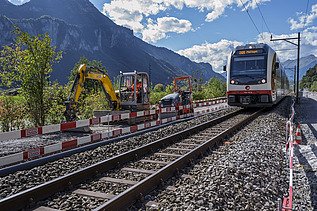 Ein Zug fährt auf einem Gleis durch eine Baustelle, rechts und links stehen Baumaschinen und Absperrungen. Im Hintergrund erheben sich hohe Berge unter blauem Himmel.
