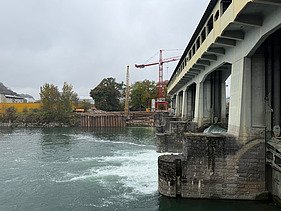 Baustelle am Flussufer: Sicht vom Wasser auf eine Staumauer, roten Kran und Spundwände der Baugrube.
