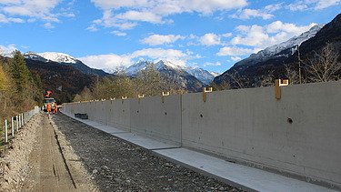 Eine lange, betonierte Stützmauer verläuft entlang einer Strasse. Daneben befindet sich ein vorbereiteter Kiesweg, links liegen Schotter und Material. Im Hintergrund erheben sich schneebedeckte Berge unter einem blauen Himmel mit Wolken.