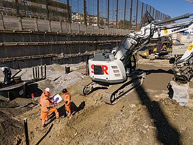 Rüschlikon Bahnhof Süd – STRABAG Tiefbau und Spezialtiefbau Drohnenaufnahme der Baugrube von vorne, zu sehen sind mehrere Bagger, im Vordergrund Bauarbeiter bei Vermessungen die zusammen in einem Kreis stehen