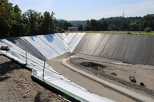Blick auf eine grosse, asphaltierte Steilböschung mit hellem und dunklem Belag unten verläuft ein Baustellenweg, im Hintergrund Wald und Landschaft.