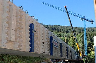 Ein langer Holzträger mit STRABAG Logo liegt auf einer Transportvorrichtung im Hintergrund heben zwei Kräne den Träger vor bewaldeten Hügeln an.