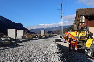 Mehrere Arbeiter in orangefarbener Schutzkleidung stehen auf einer frisch geschotterten Strasse. Eine gelbe Baumaschine steht direkt daneben. Rechts sind Wohnhäuser zu sehen.