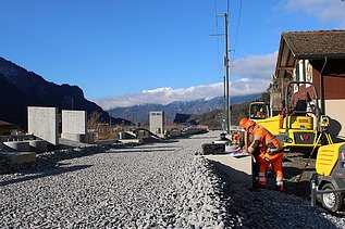 Mehrere Arbeiter in orangefarbener Schutzkleidung stehen auf einer frisch geschotterten Strasse. Eine gelbe Baumaschine steht direkt daneben. Rechts sind Wohnhäuser zu sehen.