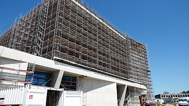 Nahaufnahme eines grossen, eingerüsteten Gebäudes auf einer Baustelle. Die Fassade ist vollständig von Gerüsten umgeben, im Vordergrund stehen ein geöffneter STRABAG Baucontainer, Baupaletten und eine Baumaschine. Rechts im Bild ein weisser Lieferwagen und ein im Bau befindliches Nebengebäude, darüber ein klarer blauer Himmel.