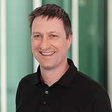 Technischer Gruppenleiter mit schwarzem Shirt lächelt in die Kamera. Im Hintergrund ist ein Lift zu sehen.