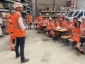 Lernende in orangefarbener STRABAG Arbeitskleidung sitzen auf Festbänken und hören zwei Personen zu, die vor ihnen sprechen