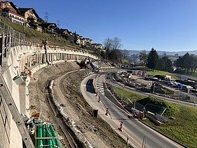  Panoramablick auf eine grosse Baustelle in Küssnacht. Eine Bohrpfahlwand stützt einen Hang mit Wohnhäusern und sichert die danebenliegende Strasse. Im Vordergrund sind Erdbewegungen und Baumaschinen zu sehen.