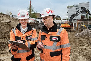 Frau mit Helm und Baustellenkleidung erklärt Mann die Arbeit