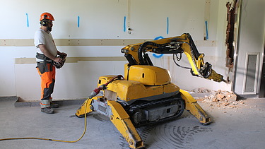 STRABAG Vorarbeiter bedient einen gelben Abbruchroboter mit Abbruchhammer im zukünftigen Dialysebereich mit ersten Einbrüchen an der Wand.