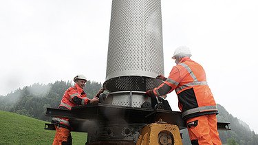 Zwei STRABAG Bauarbeiter in oranger Arbeitskleidung richten zylindrischen Filterbrunnen aus. Im Hintergrund sind grüne, bewaldete Berge zu sehen.