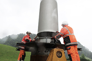 Zwei STRABAG Bauarbeiter in oranger Arbeitskleidung richten zylindrischen Filterbrunnen aus. Im Hintergrund sind grüne, bewaldete Berge zu sehen.